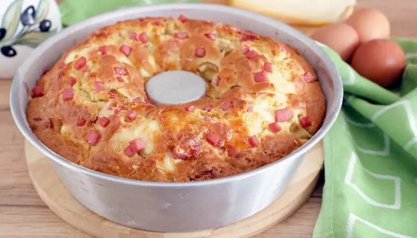 Gâteau salé végétarien doré, parsemé de dés de jambon et de fromage, dans un moule à savarin. Des œufs et du fromage sont visibles en arrière-plan.