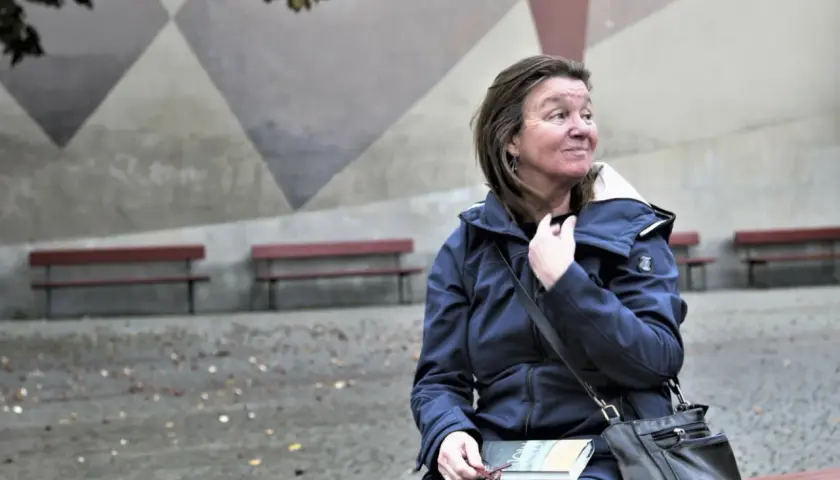 Elisabeth Rynell sitter på en bänk med en bok i knät, blickar ut mot en geometrisk vägg.