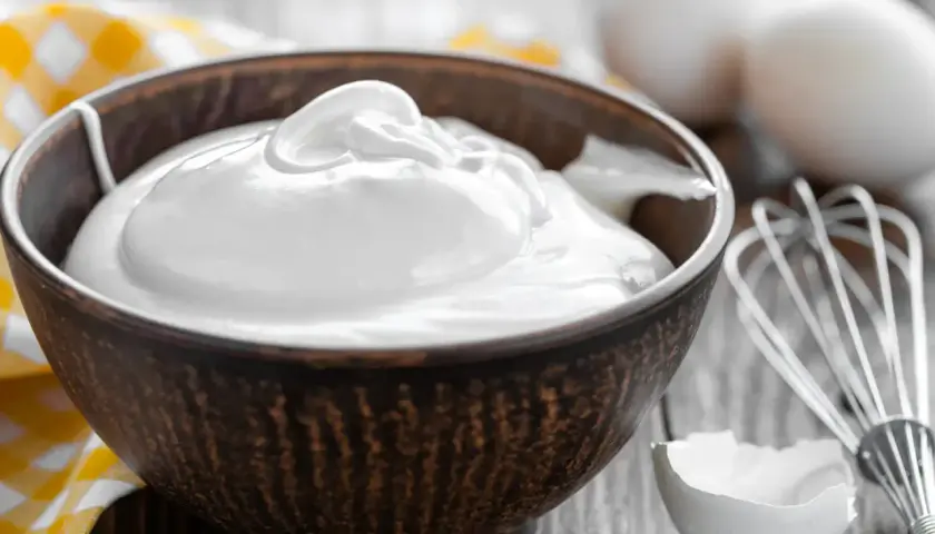 Un bol rempli de crème fouettée, blanc en neige, prêt à être utilisé pour une pâtisserie. Des œufs et un fouet sont visibles à proximité.