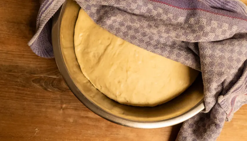 Ein goldgelber Pizzateig ruht in einer Schüssel, bedeckt mit einem karierten Tuch. Seine weiche, elastische pizzateig konsistenz verspricht eine köstliche Pizza.