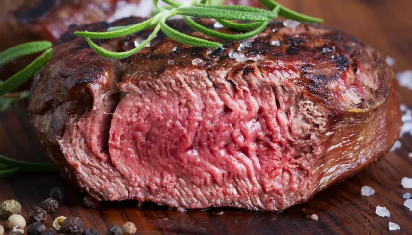 Succulenta tagliata di manzo al sangue, guarnita con rosmarino fresco e grani di pepe su un tagliere di legno.