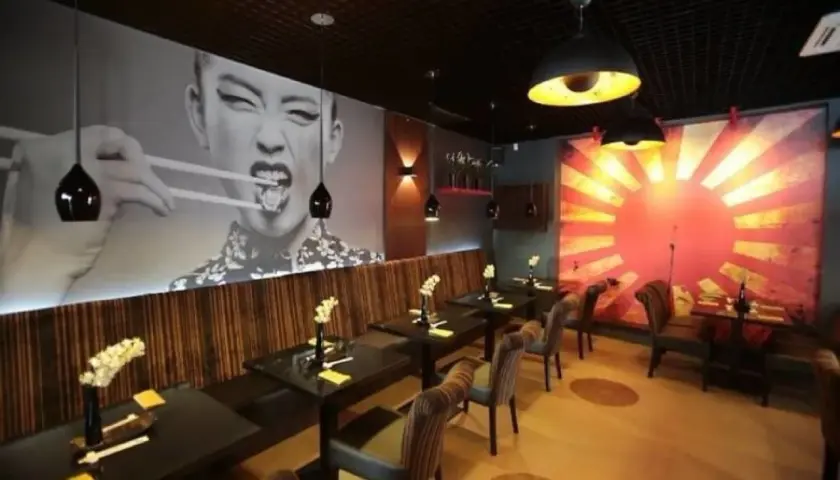 Szukasz miejsca, gdzie zjeść w Szczecinie? Wnętrze restauracji z azjatyckim klimatem, wielkoformatowym zdjęciem kobiety jedzącej pałeczkami i grafiką wschodzącego słońca.