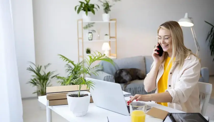 Uśmiechnięta kobieta pracuje przy laptopie i rozmawia przez telefon w domowym biurze.