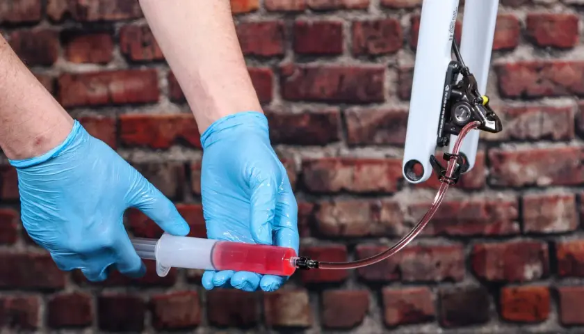 Hands in blue gloves bleed bike brakes, using a syringe filled with red fluid. This shows how much brake fluid to bleed brakes for optimal performance.