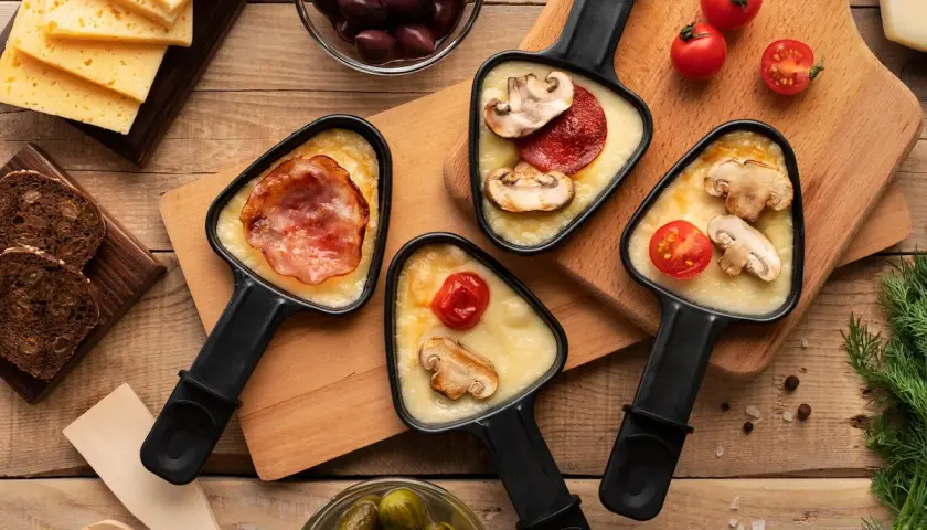Raclette party with melted cheese, bacon, mushrooms, and tomatoes. Discover par quoi remplacer le fromage à raclette pour varier les plaisirs !