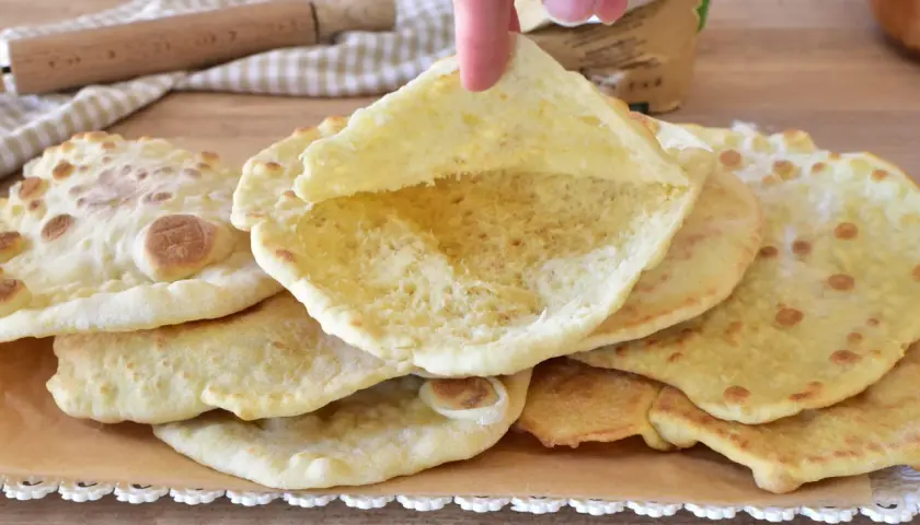 Un dito solleva un soffice pane in padella, mostrando la sua interna alveolata. Altri dischi dorati riposano su carta forno.