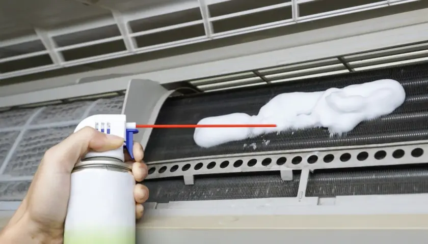 Mano rociando espuma limpiadora en las aletas de un aire acondicionado para limpiar bandeja aire acondicionado.