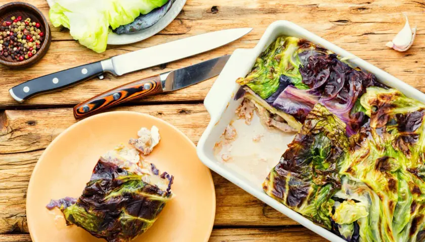 Pieczona kapusta w naczyniu żaroodpornym, obok talerz z porcją i przyprawy.