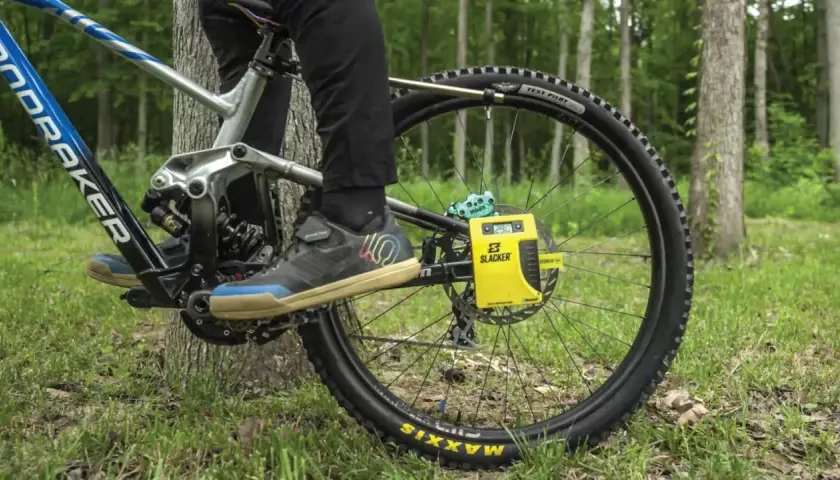 A person uses a yellow Slacker digital scale for rear shock measurements on a mountain bike, with a forest background.
