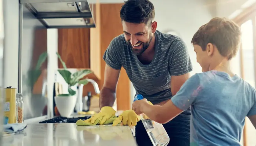 Padre e hijo ríen mientras aprenden como limpiar tu casa. El niño rocía la encimera mientras el padre la limpia con un paño.
