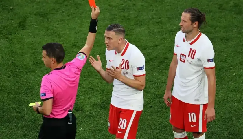 Sędzia pokazuje czerwoną kartkę, która wyklucza zawodnika z gry. Piłkarz z numerem 20 protestuje, a obok stoi jego kolega z numerem 10.