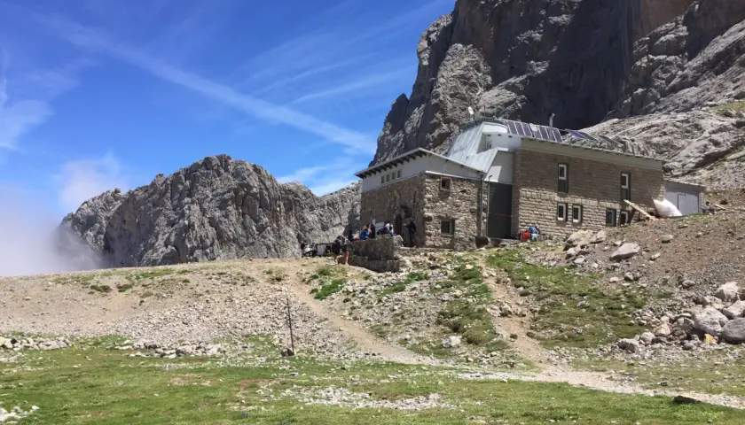 Schronisko pod masywnym **Naranjo de Bulnes**. Grupa turystów odpoczywa na tle skalistych szczytów i błękitnego nieba.