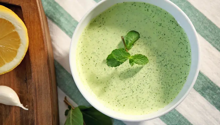 Un bol de sauce à la menthe fraîchement préparée, garni de feuilles de menthe. À côté, une tranche de citron et une gousse d'ail sur une planche en bois.