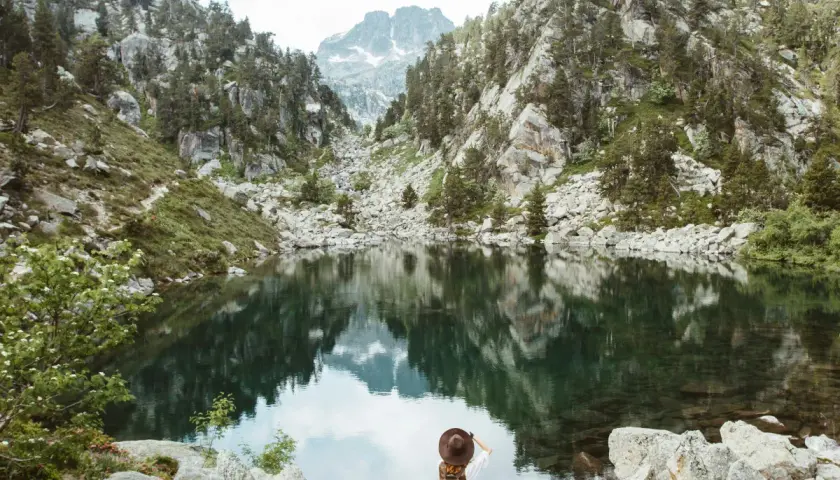 Una excursionista admira un lago sereno en el **parque nacional de los Pirineos**, rodeado de montañas rocosas y pinos.