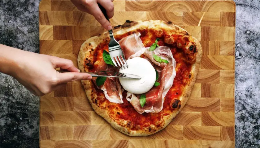 Une pizza burrata avec du prosciutto et du basilic, prête à être dégustée. Des mains coupent la burrata crémeuse.
