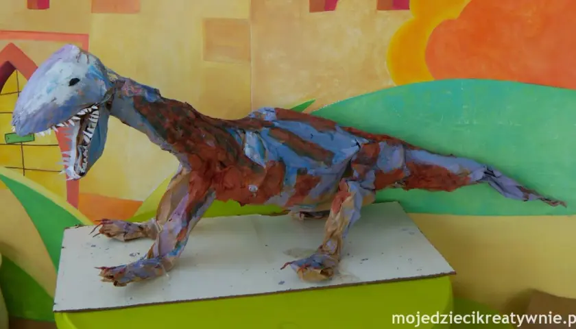 Kolorowa dinozaury praca plastyczna z papieru, z otwartą paszczą i ostrymi zębami, na tle malowanej ściany.