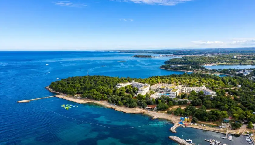 Hotel Delfin Plava Laguna z lotu ptaka, otoczony zielenią i błękitnym morzem. Widoczne baseny, plaża i przystań z łodziami.