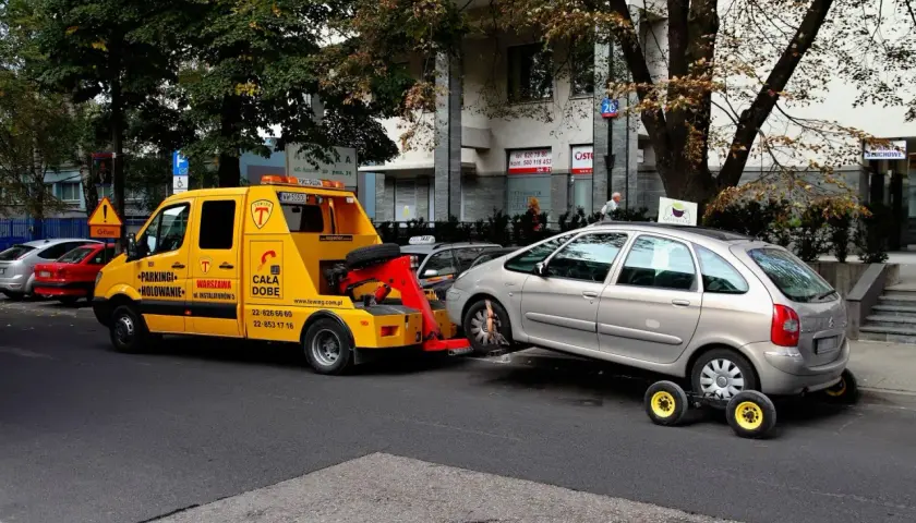 Żółty holownik odholowuje srebrny samochód. Kierowca dostał mandat za parkowanie.