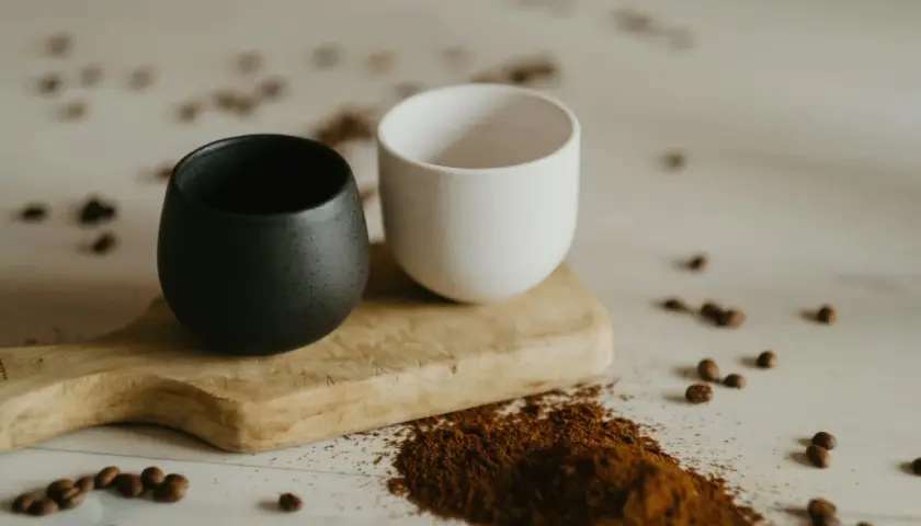 Dwa ceramiczne kubki, czarny i biały, na drewnianej desce. Obok rozsypana mielona kawa, która sugeruje, czy kawe sypana sie miesza.