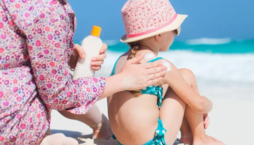 Mamma applica la crema solare a una bambina sulla spiaggia, prevenendo la dermatite da sabbia.