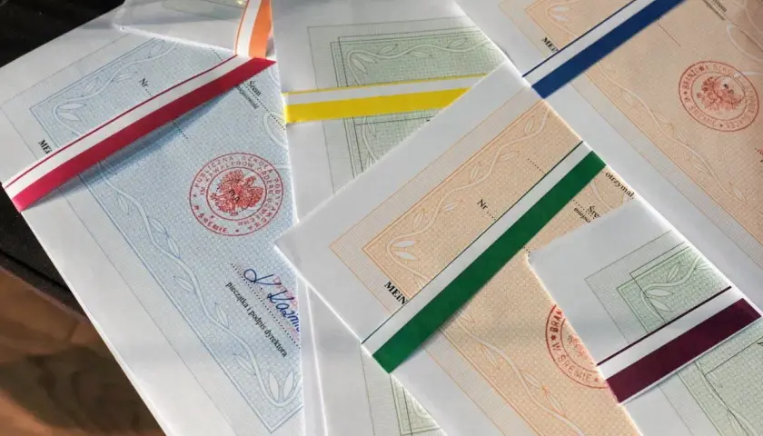 Stos papierów z kolorowymi wstążkami, każdy z pieczęcią i podpisem dyrektora. To świadectwo ukończenia szkoły.