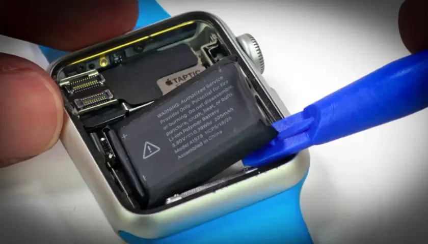 Wymiana baterii Apple Watch: wyjmowanie starego ogniwa za pomocą niebieskiego narzędzia.