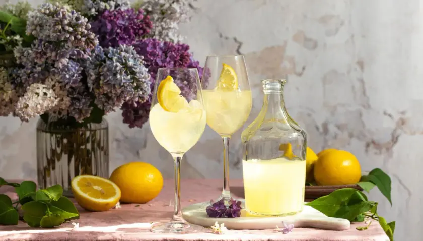 Erfrischendes Lemon Spritz Rezept: Zwei Gläser mit Eis und Zitronenscheiben, daneben eine Karaffe mit Zitronenlikör und frische Zitronen. Ein Blumenstrauß aus Flieder im Hintergrund.