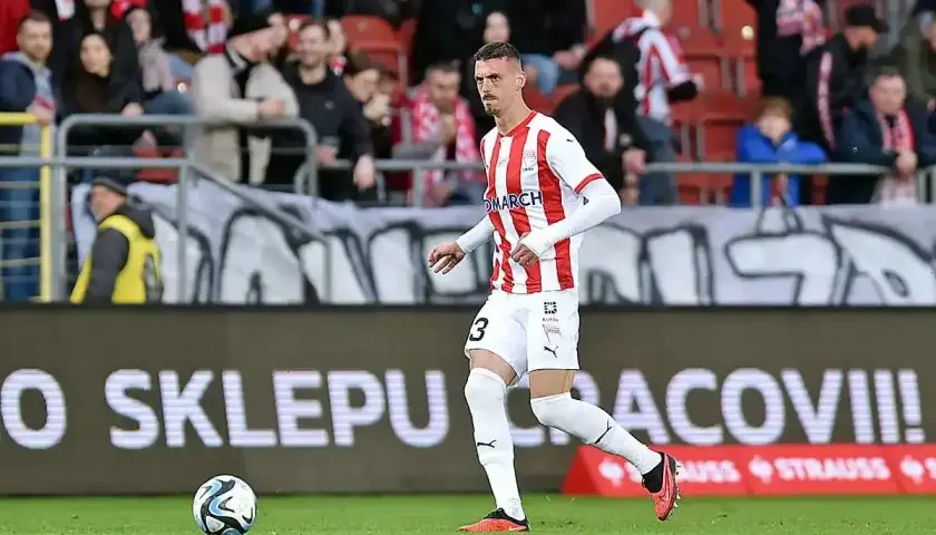 Piłkarz w koszulce z numerem 3 biegnie z piłką. W tle kibice i baner "O SKLEPU CRACOVII!".