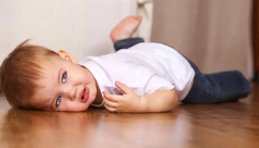 Un bebé de 16 meses con ojos azules y cabello castaño yace en el suelo de madera, vestido con una camiseta blanca y jeans.