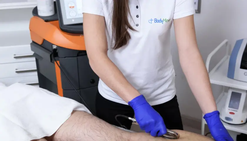 Fizjoterapeuta w niebieskich rękawiczkach wykonuje zabieg na nodze pacjenta za pomocą urządzenia z elektrodą w kształcie gęsiej stopki.