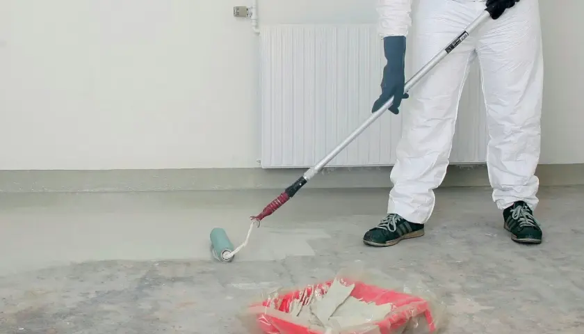 Person i skyddsdräkt målar betonggolv med roller. En färgtråg med ljus färg står på golvet.