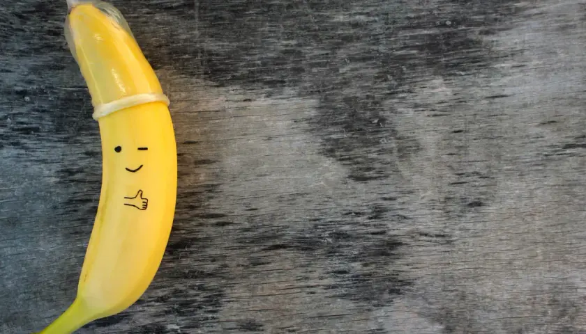 Banan z namalowaną buźką i kciukiem w górę, ubrany w **kondom**. To zabawne przypomnienie o bezpieczeństwie.