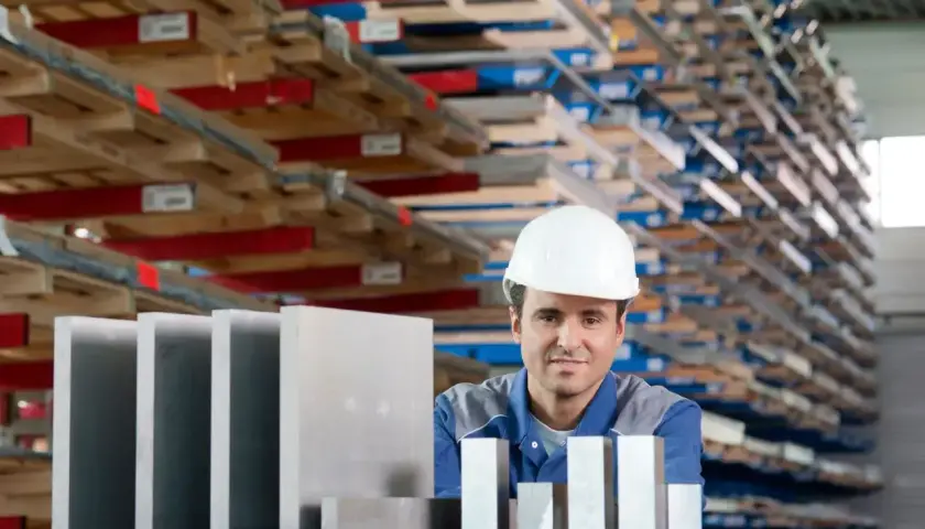 Pracownik w białym kasku stoi przed regałami pełnymi materiałów, obok niego leżą różne stopy aluminium.