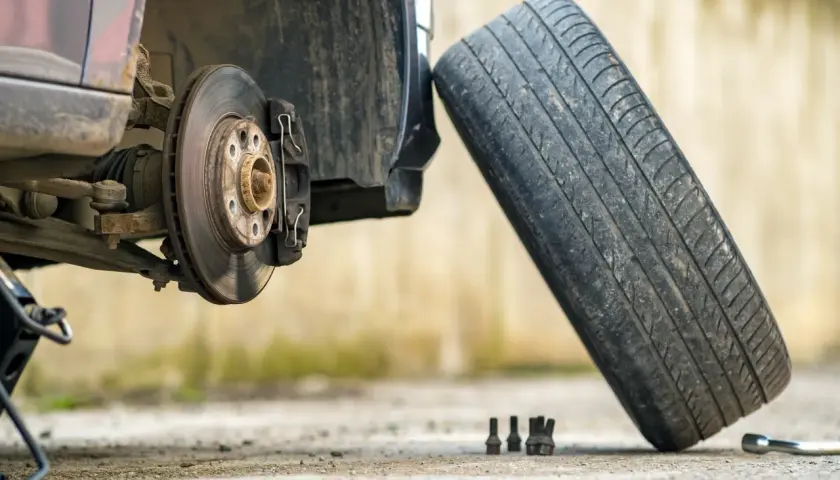 Un coche con el disco de freno expuesto y un neumático viejo apoyado. Se ven tuercas y una llave inglesa en el suelo.