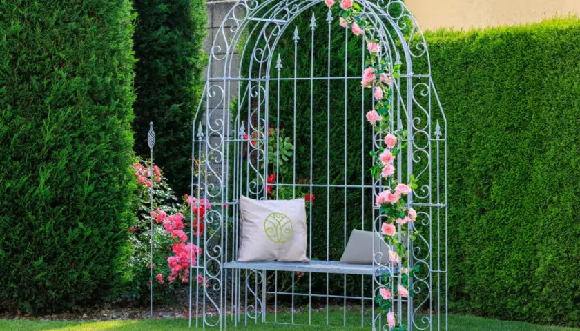 Ein weißer Rosenbogen mit rosa Rosen, Sitzbank und Kissen. Tolle Rosenbogen Gestaltungsideen für Ihren Garten.