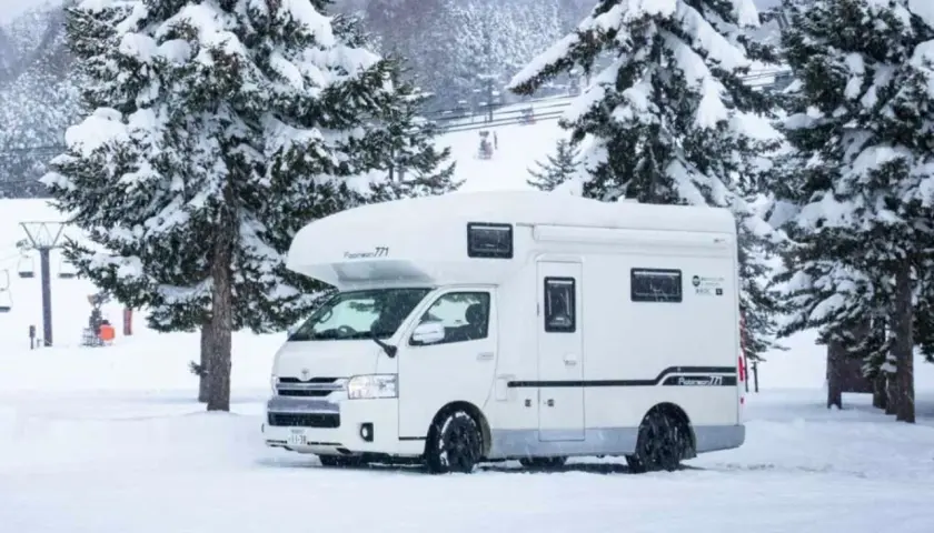 Camping car japonais blanc garé dans la neige, entouré de sapins enneigés. Des remontées mécaniques sont visibles en arrière-plan.