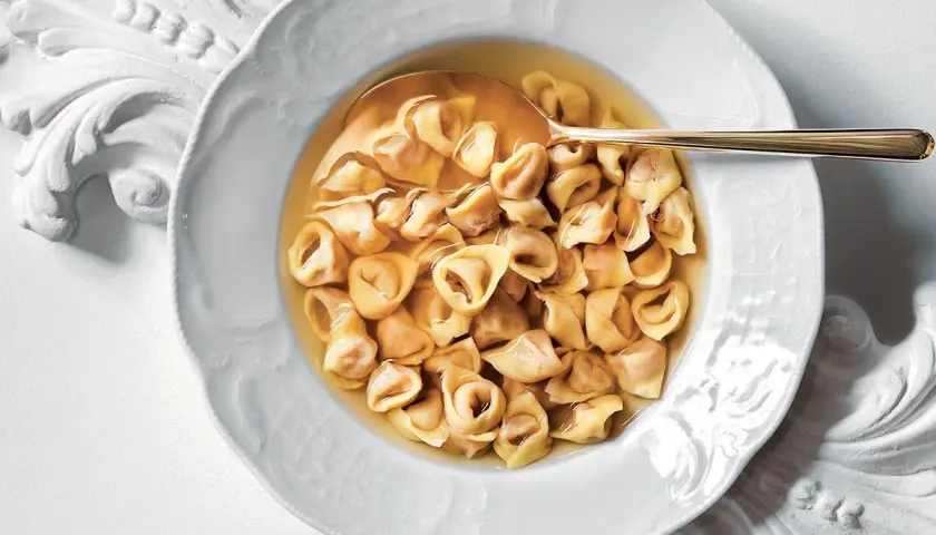 Un piatto di tortellini in brodo, un classico confortante, servito in una ciotola bianca decorata con un cucchiaio dorato.