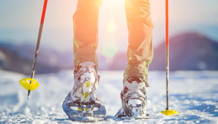 Piernas con raquetas de nieve y bastones listos para excursiones en la nieve bajo un sol brillante.