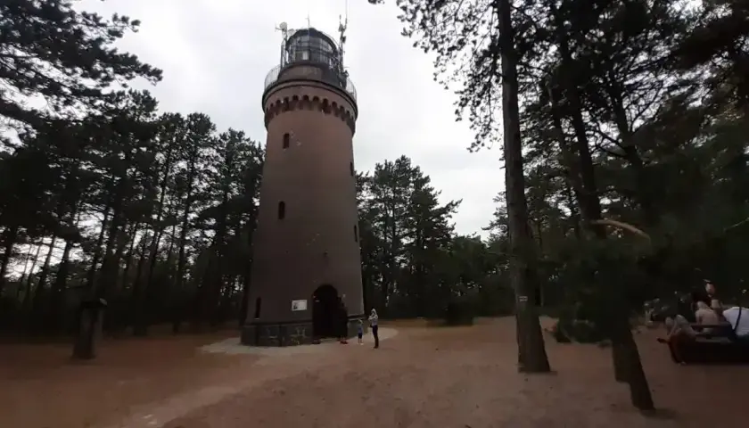 Latarnia morska w Czołpinie, otoczona sosnowym lasem, góruje nad piaszczystym terenem. W oddali widać fragmenty wydm.
