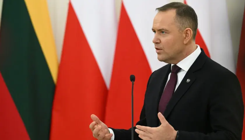 Karol Nawrocki, w garniturze, przemawia na tle flag. Jego gestykulacja i pewność siebie mogą być powodem, dlaczego Karol Nawrocki stał się nowym liderem sondaży zaufania na prawicy.