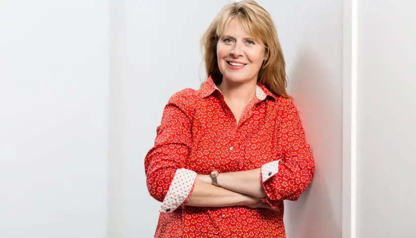 Petra Weber, eine lächelnde Frau mit blonden Haaren, trägt ein rotes Hemd mit Blumenmuster und hat die Arme verschränkt.