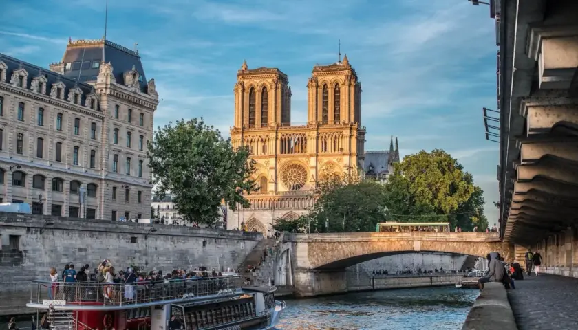 Katedra Notre Dame w Paryżu nad Sekwaną, z łodzią turystyczną i mostem w tle.
