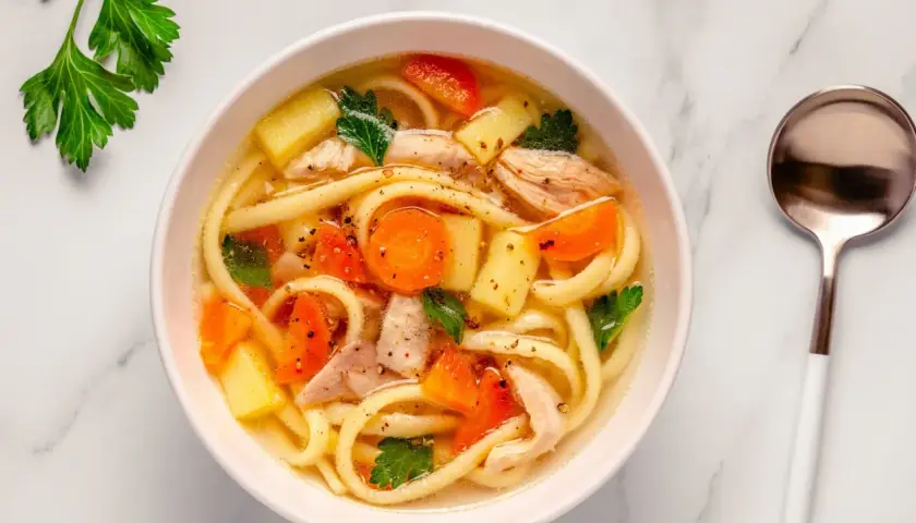 A miseczce paruje aromatyczna zupa z makaronem, z kawałkami kurczaka, marchewki i ziemniaków, ozdobiona natką pietruszki.