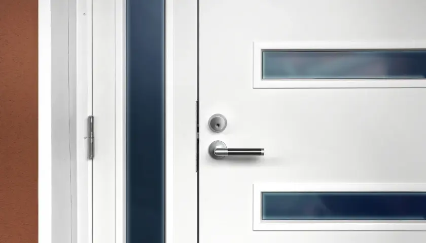 Nowoczesne białe drzwi wejściowe z metalową klamką i wąskimi, ciemnymi przeszkleniami. Dowiedz się, jak zmierzyć drzwi wejściowe.