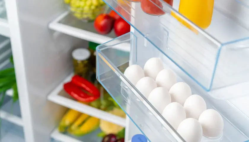 Ein Kühlschrank voller frischer Lebensmittel. In der Eierablage liegen viele weiße Eier, die noch gut sind. Daneben sind rote Paprika, Maiskolben und Trauben zu sehen.