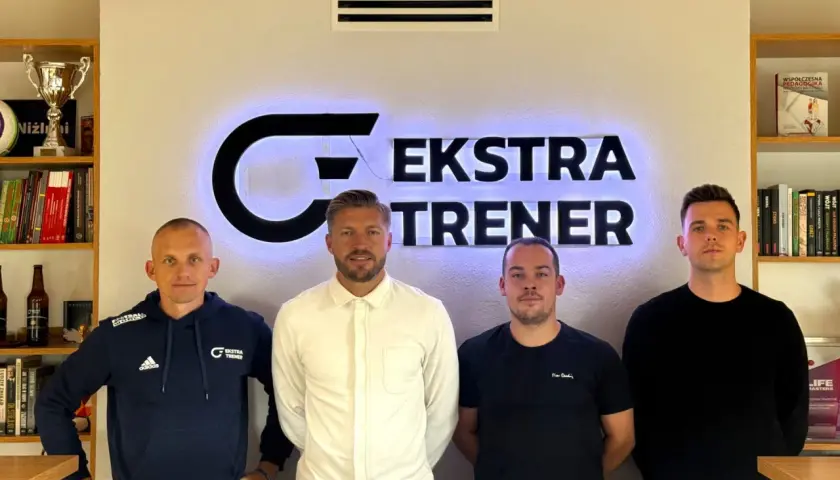 Czterech mężczyzn stoi przed podświetlanym logo "EKSTRA TRENER". Dwóch z nich to srodkowy pomocnik, który jest trenerem.