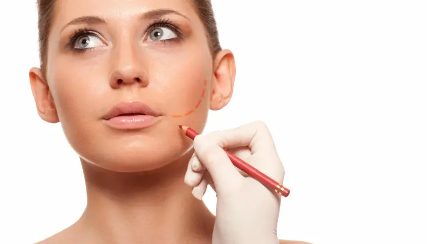 En hand i handske markerar ansiktet med en röd penna, förberedelse för buccal fat removal.