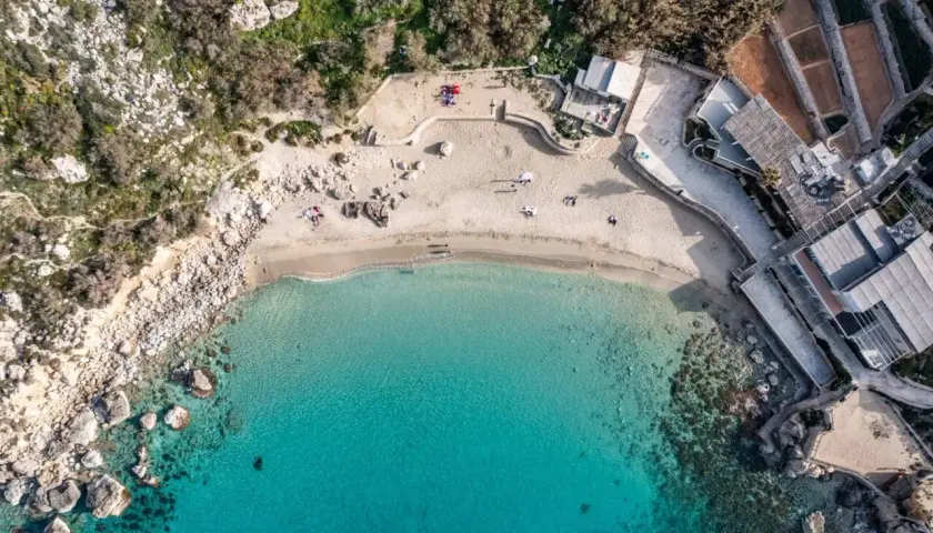 Widok z lotu ptaka na rajską zatokę na Malcie, z turkusową wodą, piaszczystą plażą i budynkami przy brzegu.