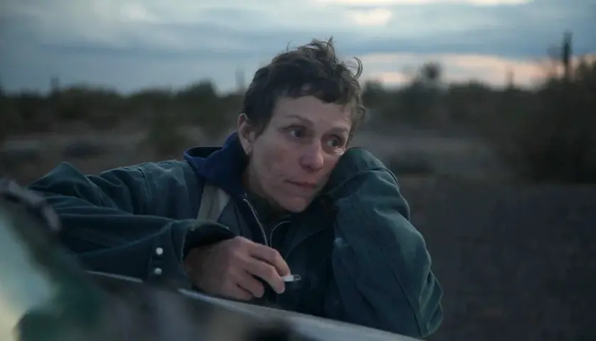 Skådespelare Frances McDormand lutar sig mot en bil i ökenlandskap, med en cigarett i handen.