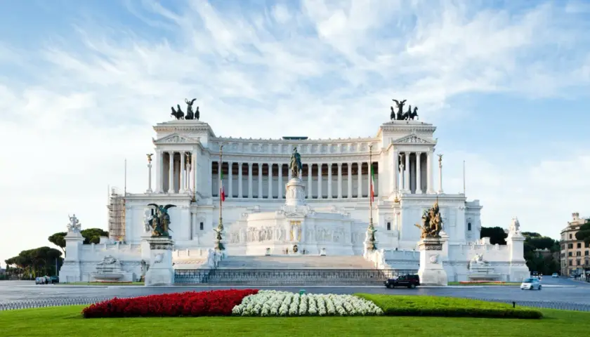 Il Vittoriano, monumento imponente, si erge maestoso. Vicino alla fermata metro Altare della Patria, un prato verde e fiori rossi e bianchi creano un contrasto vibrante.